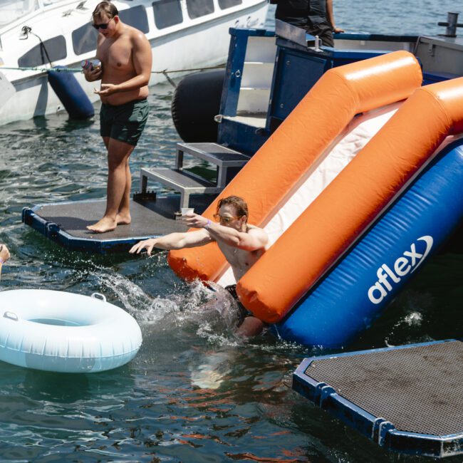 A person slides down an inflatable water slide into a lake from a dock. Two people stand nearby, one holding a drink. Boats are anchored in the background under a sunny sky.