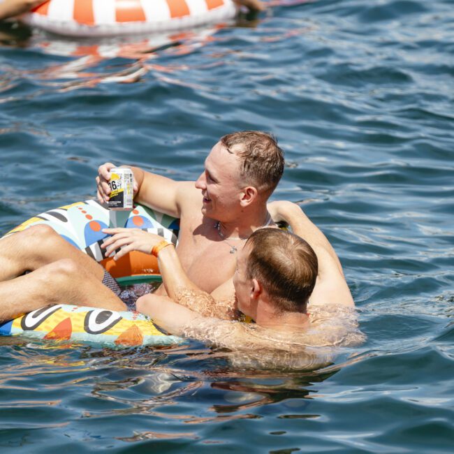 Two people are lounging on a colorful inflatable ring in a body of water. One is holding a can of drink. The sun is shining, creating a bright and cheerful atmosphere as they relax and enjoy the moment.