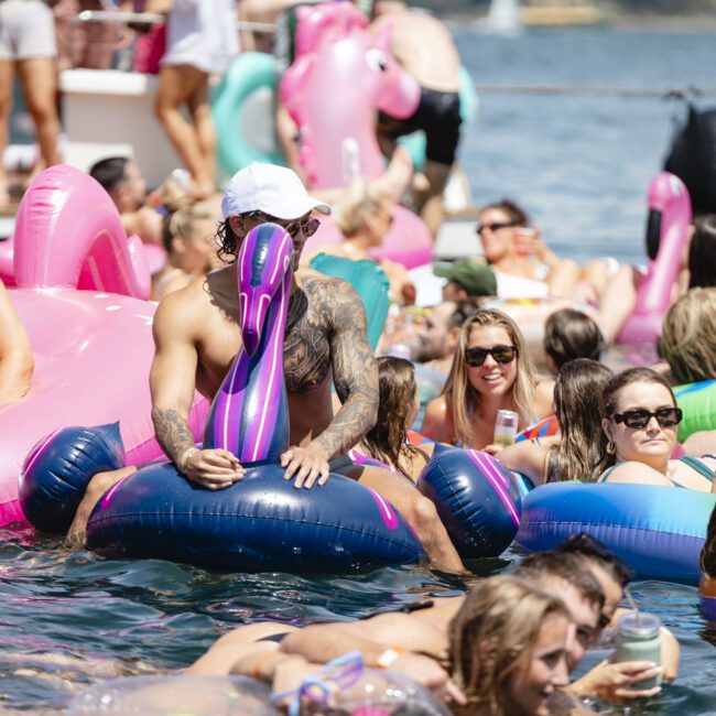 A group of people enjoying a sunny day in the water on colorful inflatable pool floats shaped like flamingos and unicorns. The scene is lively with many people relaxing and socializing in their swimsuits.