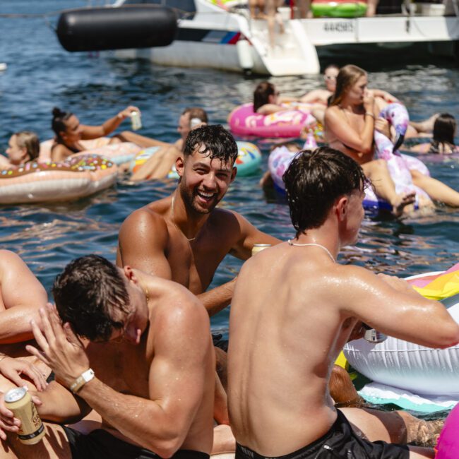 A group of people enjoying a sunny day on a boat and in the water. Some are sitting on the boat's edge, holding drinks, while others float on colorful inflatables in the water. A yacht is visible in the background. Everyone appears to be having fun.