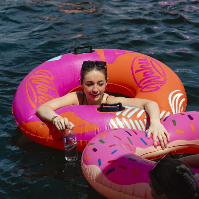 A person relaxes in a colorful donut-shaped pool float on the water, holding a bottle. Other people on floats are visible in the background. The scene is sunny and festive.