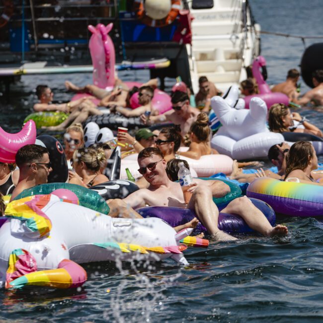 A group of people enjoying a sunny day on the water, lounging on colorful inflatable floats near a boat. Some are in the water, surrounded by friends and more inflatables, creating a vibrant and lively atmosphere.