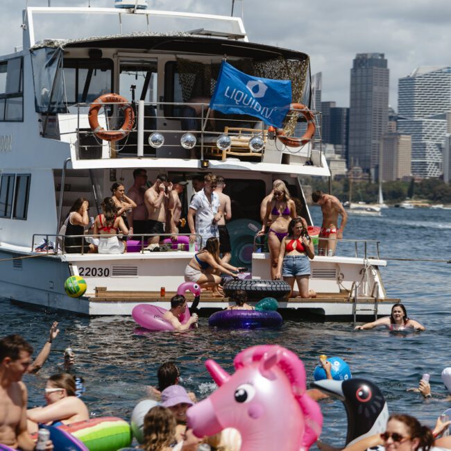 A group of people enjoying a party on a yacht and in the water, surrounded by inflatable floats like a unicorn and donuts. The city skyline and blue sky are visible in the background.