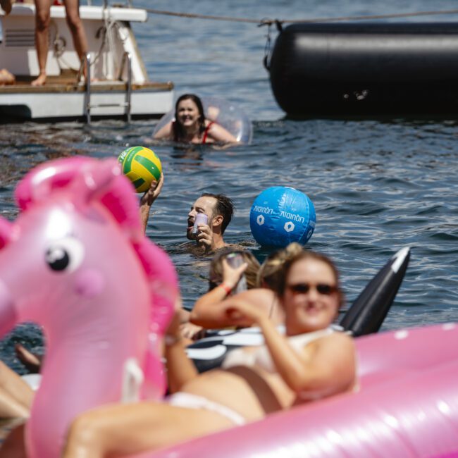 People enjoying a sunny day on the water, with some floating on inflatable toys, including a pink unicorn. A person in a mask is seen holding a water ball. A boat and an inflatable platform are in the background.