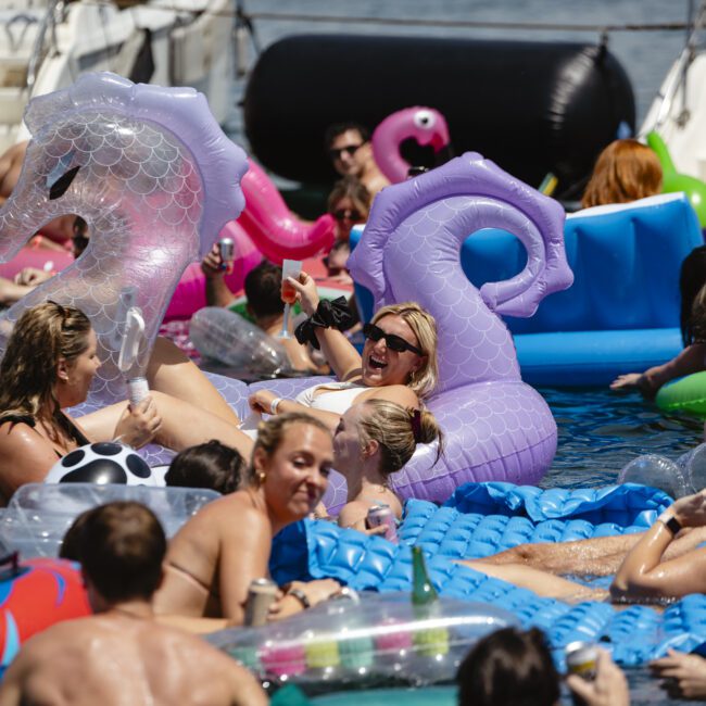 A lively pool party with people relaxing on inflatable floats in various shapes, including dragons and swans. The atmosphere is festive, with attendees enjoying drinks and the sun. Boat visible in the background.