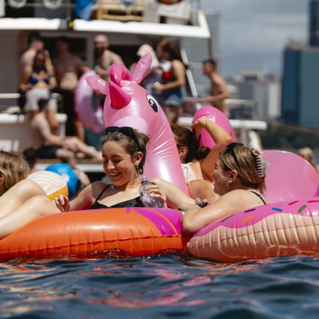 A group of people relaxing on colorful inflatable floaties, including a pink unicorn and donuts, in a body of water. A boat and city skyline are visible in the background. It appears to be a sunny day with many people enjoying the scene.
