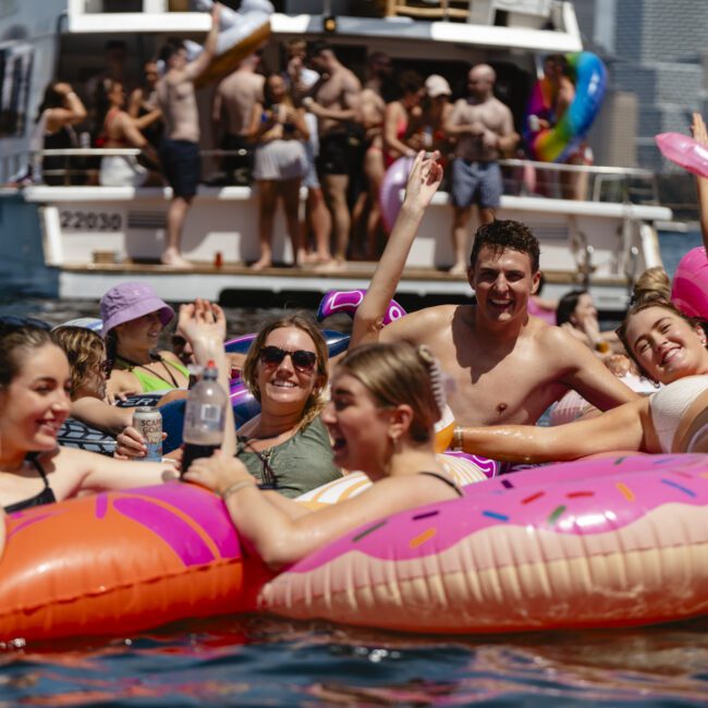 A group of people is enjoying a sunny day in the water, sitting on pink donut and unicorn inflatables. A boat with more people is in the background, along with city buildings. Everyone appears to be having fun and socializing.