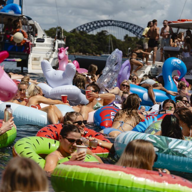 A vibrant scene of people lounging on inflatable pool floats in a sunny body of water. The floats are shaped like unicorns, watermelons, and more. Several boats and a bridge are in the background. Everyone is enjoying a lively social event.