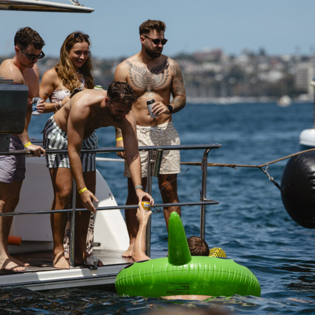 A group of people on a boat are watching as a person in a green inflatable tube floats in the water. One person is bending over to help. The background features a clear blue sky and distant shoreline.