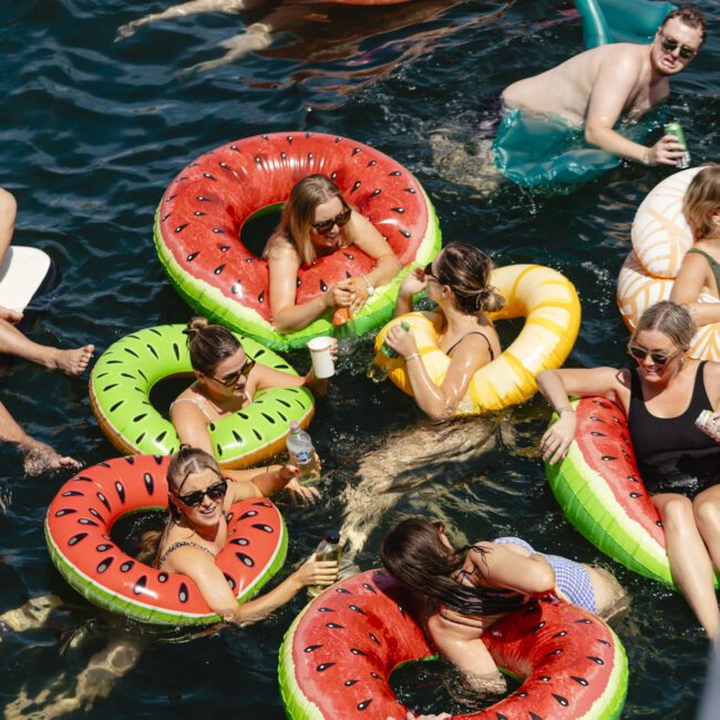 A group of people relaxing on colorful inflatable floats shaped like donuts and watermelons in a body of water. They are enjoying the sunny weather, holding drinks, and engaging in conversation. The atmosphere is lively and cheerful.