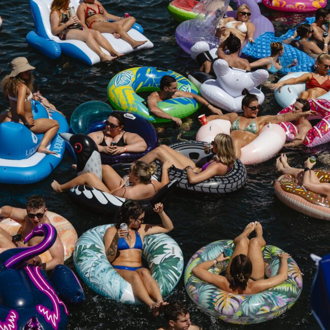 A crowded group of people relaxing on colorful inflatable pool floats of various shapes, like donuts and flamingos, in a body of water. The scene is lively and sunny, with many enjoying the summer day.