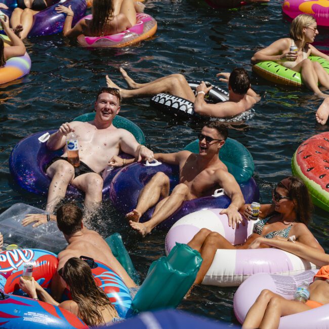 A lively scene of people floating on colorful inflatable tubes in a pool. Some are holding drinks, chatting, and enjoying the sun. The tubes are shaped like donuts and other fun designs, creating a festive, relaxed atmosphere.