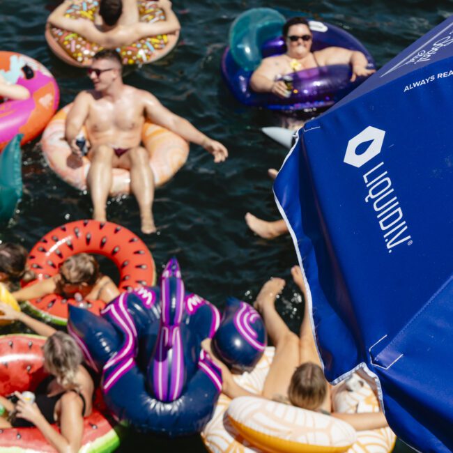 People relaxing on colorful inflatable floats in a body of water. They are socializing under the sun. A blue umbrella with text and a logo partially visible on the right.