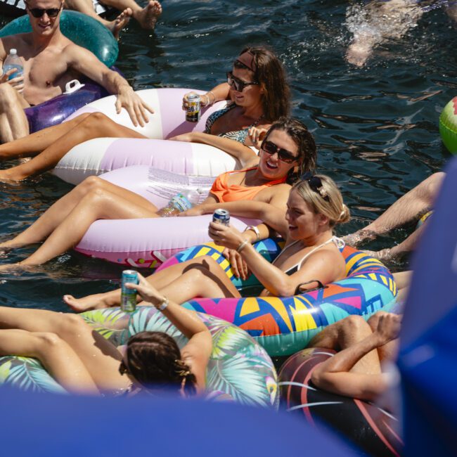 A group of people relaxing on colorful inflatable floats in the water, holding drinks. The scene is lively, with some wearing sunglasses and swimwear. A blue triangular banner with the text "Yacht Social Club" is partially visible at the bottom.