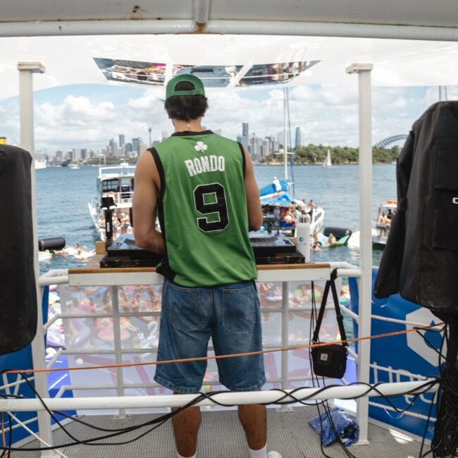 A person wearing a green "Ronaldo 9" jersey and a green cap is DJing on a boat. Large speakers are on either side. A crowd is dancing on deck, with a city's skyline visible across the water under a blue sky.