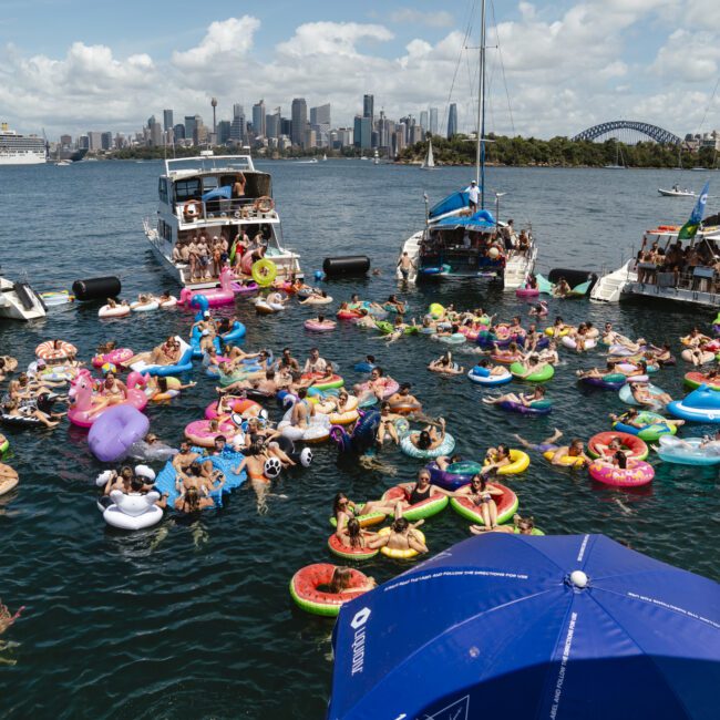 A lively scene on the water with people on inflatable floats and boats, enjoying a sunny day. The background features a city skyline and the Sydney Harbour Bridge. The atmosphere is festive with many people in swimsuits relaxing and socializing.