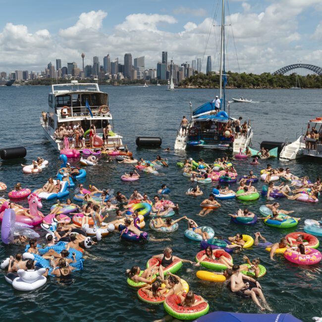 Crowd of people on colorful inflatable rings enjoying a sunny day on the water near docked boats. Sydney's skyline and Harbour Bridge are visible in the background.