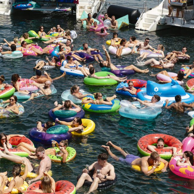 A group of people float on colorful inflatable tubes and rafts in the water. The scene is lively, with various floats, including a swan and a flamingo. Boats are visible in the background, creating a festive atmosphere.