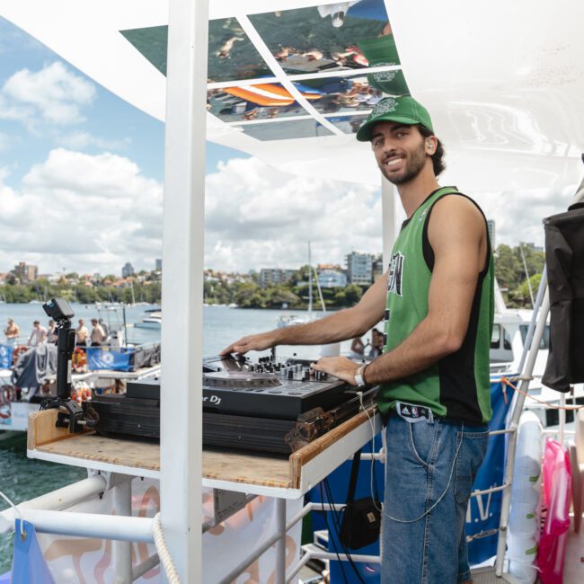 A DJ wearing a green tank top and cap is playing music on a boat deck. The background shows people enjoying themselves on nearby boats and inflatables on a sunny day, with a blue sky and clouds overhead.