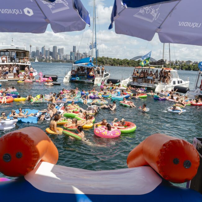 A large group of people float on colorful inflatable toys in a sunny harbor near several anchored boats. Umbrellas are on nearby decks. The city skyline is visible in the background under a partly cloudy sky.