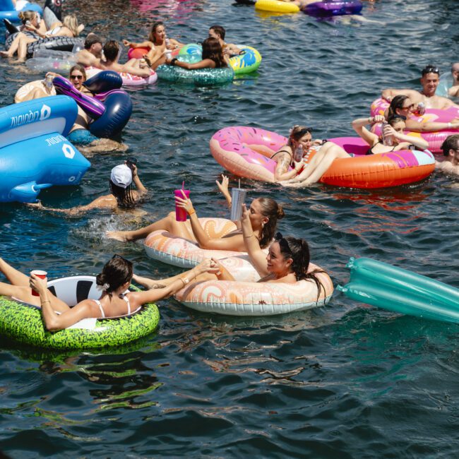 A group of people relax in colorful inflatable rafts on a sunny day in a body of water. Many hold drinks and wear swimsuits, enjoying the lively atmosphere. The scene is filled with floats in various shapes and colors, including rings and animals.