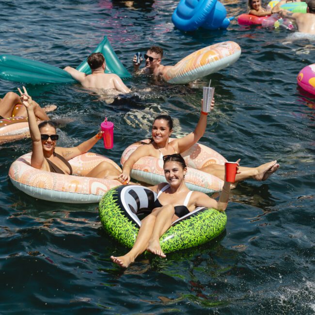 A group of people relaxing and smiling on inflatable floats in a lake. They are holding cups and enjoying sunny weather. Other colorful inflatables are scattered around them, and more people are in the background.