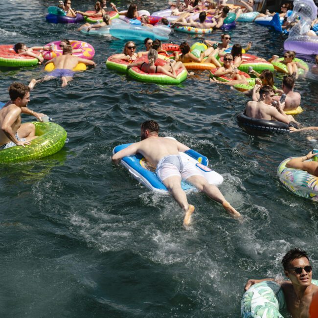People are enjoying a sunny day on a lake, floating on colorful inflatables. They are socializing and relaxing in the water. Some inflatables are shaped like animals and others are simple tubes. The atmosphere is lively and festive.