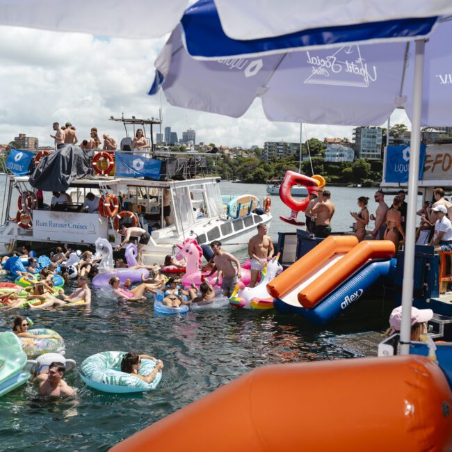 People enjoy a lively boat party on the water, surrounded by various inflatable pool floats. Boats are anchored nearby, and many people are swimming and relaxing under large umbrellas. City buildings are visible in the background.