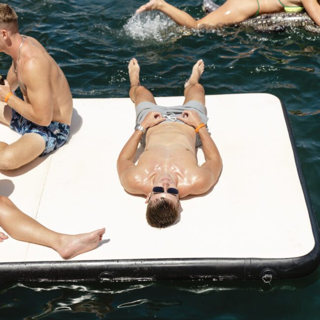 A person is lying on a floating platform on a body of water, wearing sunglasses and swim trunks. Others are sitting and swimming nearby. The scene is sunny and relaxed, ideal for a day at the lake or ocean.