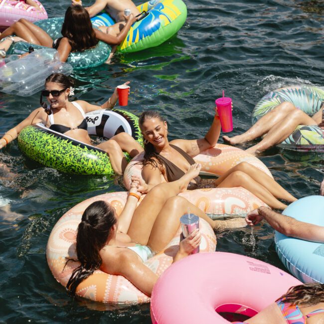 A group of people are floating on colorful inflatable rings in a body of water, enjoying a sunny day. They are smiling, talking, and relaxing. Some hold drinks, and the atmosphere is lively and festive.