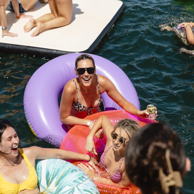 A group of people enjoying a sunny day in the water. Some are on colorful inflatable floats. One person in sunglasses holds a drink on a purple float. Others in swimsuits relax and chat. The scene is lively and joyful, set against a backdrop of water.