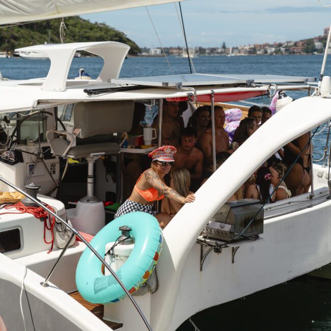 A group of people are enjoying a sunny day on a sailboat. Some are sitting under the canopy, while one person in colorful attire stands by the side. A turquoise life preserver is attached to the boat. Water and greenery are visible in the background.