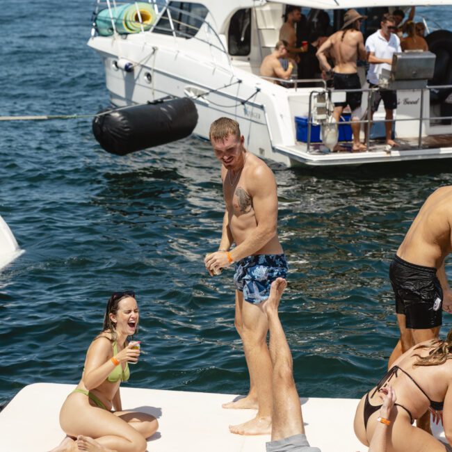 People enjoying a sunny day on the water. A woman in a bikini laughs while holding a drink. A man stands nearby, and another man is lying down, playfully splashing water. A boat with several people is in the background.