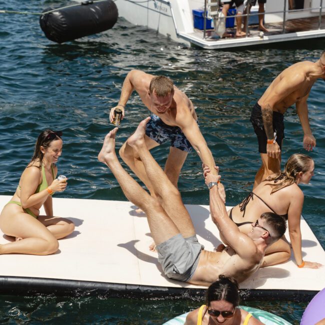 A group of people enjoy a sunny day on the water. Some are on a floating platform: one person helps another perform a balancing act. Others relax on inflatable loungers. A nearby boat has more people onboard. It’s a lively, fun scene by the water.
