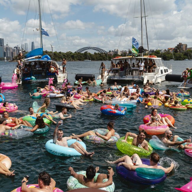 A lively crowd of people enjoying a sunny day in the water, floating on colorful inflatable tubes. Boats are nearby, and the city skyline with a bridge can be seen in the background. The atmosphere is festive and relaxed.