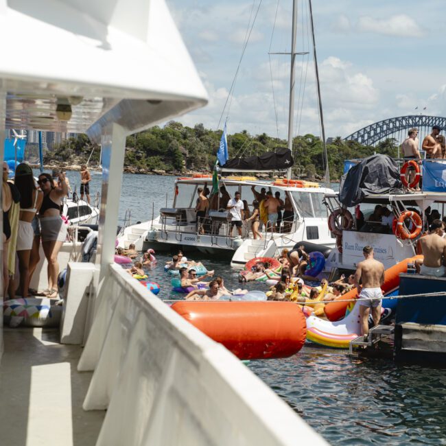 People enjoying a lively boat party on a sunny day, with several boats docked close together. Attendees relax on inflatable floats in the water, while others gather on the decks. The Sydney Harbour Bridge is visible in the background.