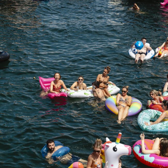 Aerial view of people enjoying a sunny day on a lake, floating on colorful inflatable rings and shapes. The water is dark blue, and the scene is lively with various floats including a unicorn and a tire.