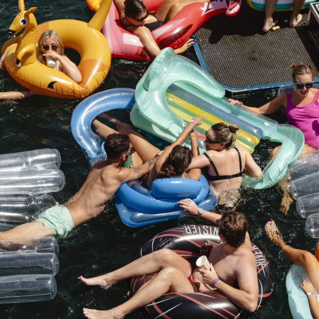 A group of people are relaxing on inflatable pool floats in the water, enjoying a sunny day. The floats are shaped like various objects, and one person is sipping a drink. The scene is vibrant, with laughter and social interactions.