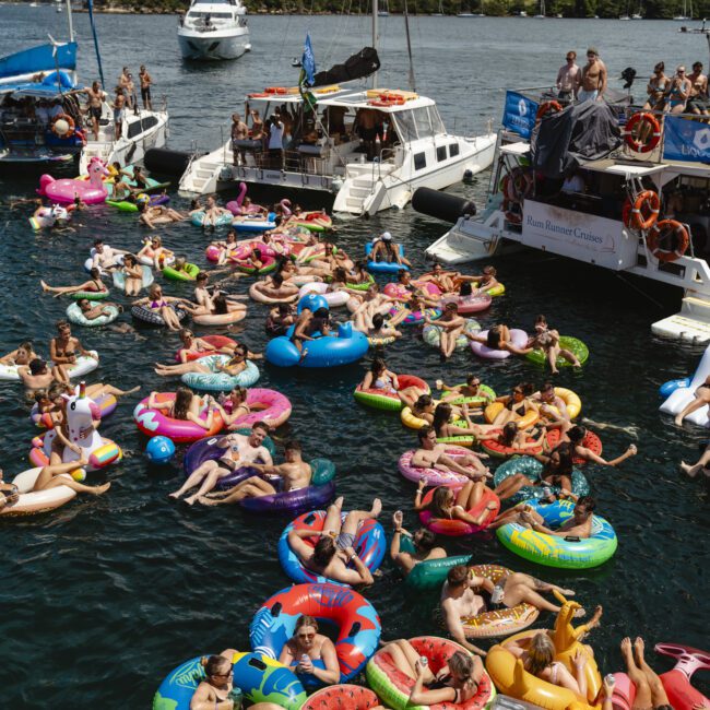 A large group of people relax on colorful inflatable floaties in a body of water. Boats are docked nearby, and a city skyline with a bridge is visible in the background. The scene is lively and festive.