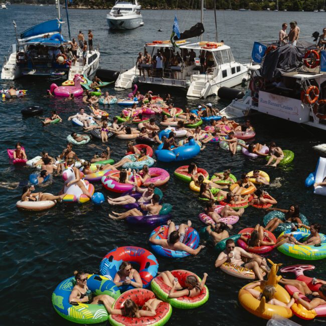 A group of people relax on colorful inflatable floats in a sunny harbor near Sydney. Boats surround the swimmers, and the Sydney skyline and iconic bridge are visible in the background. The scene is festive and crowded.
