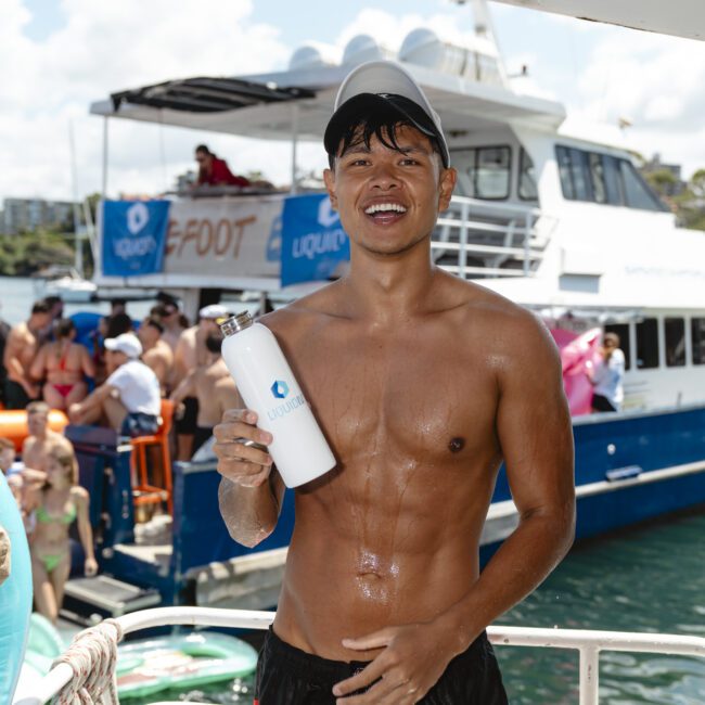 A man on a boat is holding a white water bottle and smiling at the camera. He's shirtless and wearing red and black swim shorts, with a gray cap. In the background, another boat is filled with people enjoying the sunny day on the water.