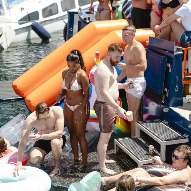 People enjoying a sunny day on a boat with inflatables in the water. The scene is lively, with individuals in swimsuits relaxing and socializing. A colorful slide and various floaties add to the festive atmosphere.