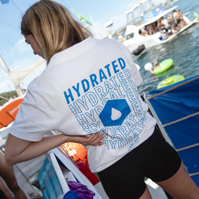 A person wearing a "Hydrated" shirt stands on a boat near water. Other people are relaxing on inflatable tubes and boats in the background. The scene is lively and sunny.