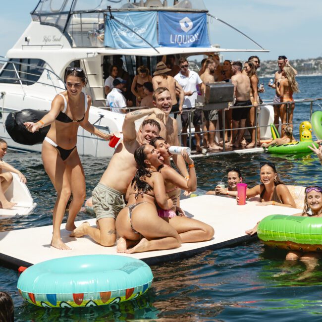 A group of people on a floating platform in the water are taking a selfie. They are surrounded by inflatable pool rings. In the background, a yacht is docked with more people on board. The sky is clear and sunny.