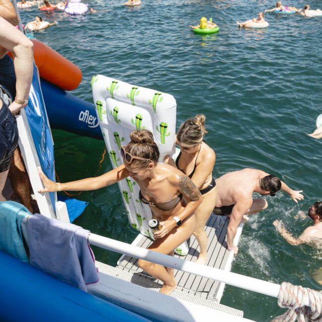 People enjoying a sunny day on a floating platform in a lake. Two women climb stairs with inflatable floats, while others swim nearby with colorful floats. Towels hang from the railing, and the scene captures the lively atmosphere of outdoor fun.