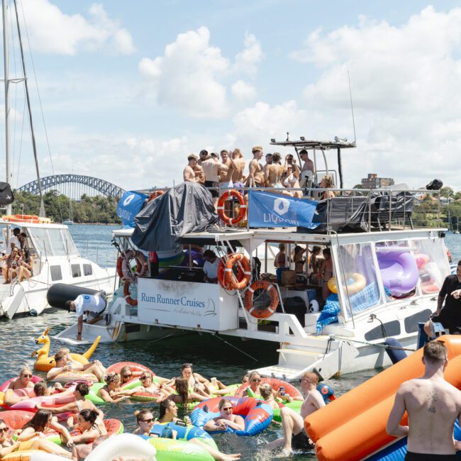 People enjoy a party on a boat filled with colorful inflatables, surrounded by others on floaties in the water. Another boat is nearby, and a bridge and cityscape are visible in the background under a partly cloudy sky.