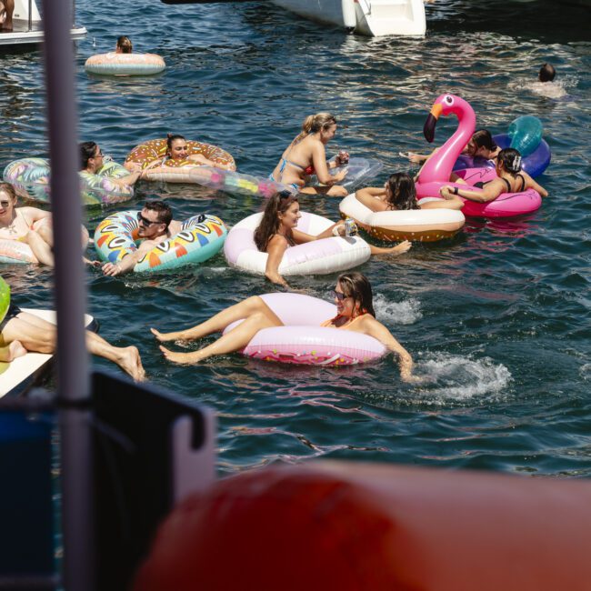 A group of people relaxing on colorful inflatable floats in the water. The floats include a pink flamingo, a donut, and a unicorn. Boats are anchored nearby. The scene is lively and sunny.