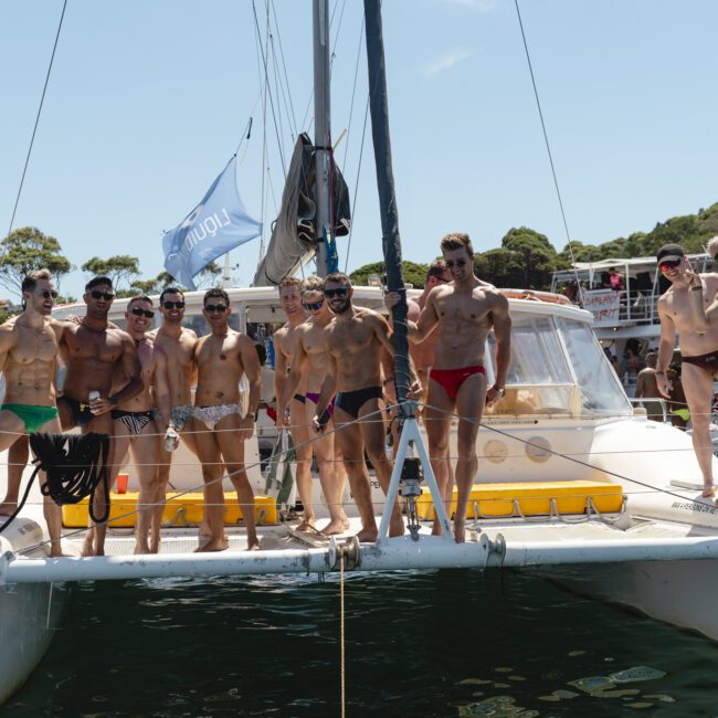 A group of people wearing swimsuits stand on the front of a catamaran in a sunny setting. There are other boats and people visible in the background, with trees and a clear blue sky overhead.