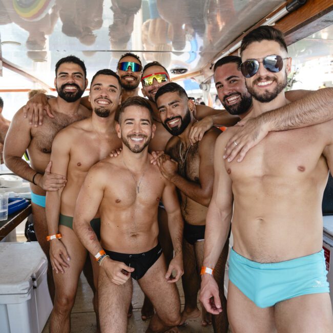 A group of seven smiling men posing together on a boat. They are wearing swim trunks and sunglasses, creating a joyful and relaxed atmosphere. The ceiling reflects some of their images, adding to the lively scene.