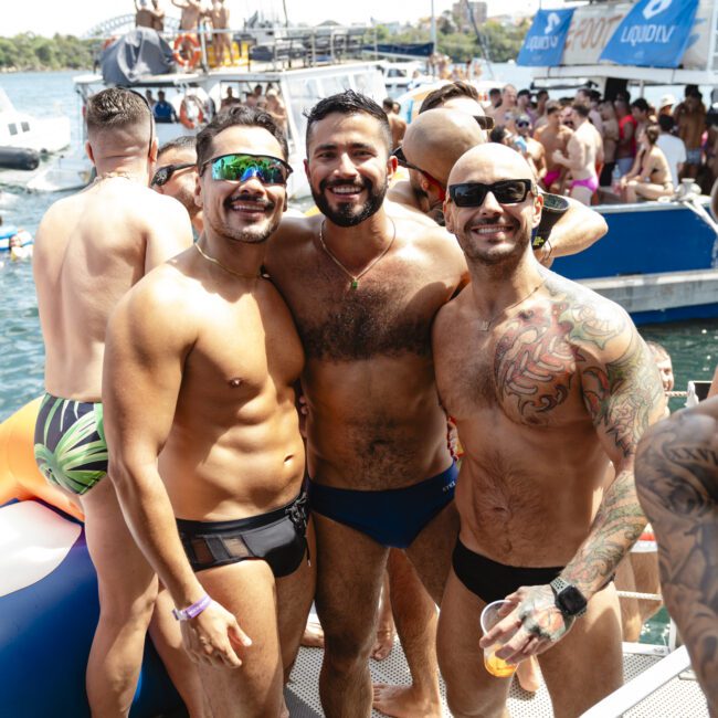 A group of four men on a boat at a lively pool party. They are wearing swim trunks, sunglasses, and have cheerful expressions. Other people are enjoying the water and surrounding boats, creating a festive atmosphere under a sunny sky.